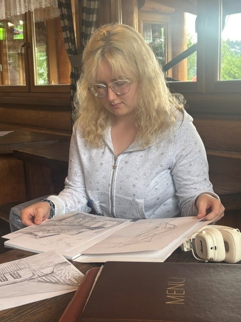 Dziewczyna w okularach siedząca przy stole we wnętrzu restauracji lub schroniska, przeglądająca album ze szkicami architektonicznymi. Obok niej leżą słuchawki.