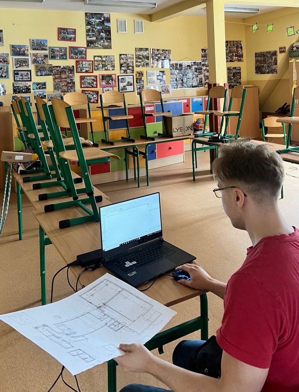 Młody mężczyzna w czerwonej koszulce siedzi przy szkolnej ławce, trzymając w ręku papierowy plan budynku i pracując na laptopie z programem do projektowania (CAD). W tle widać ułożone na stołach krzesła.