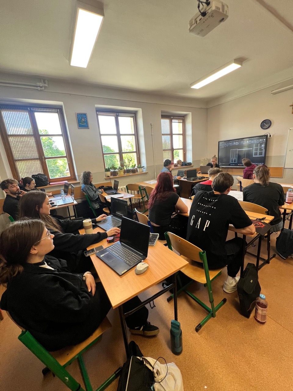 Grupa młodzieży siedzi w sali lekcyjnej przy ławkach z laptopami, uczestnicząc w zajęciach. Na dużym ekranie z przodu wyświetlony jest projekt architektoniczny.