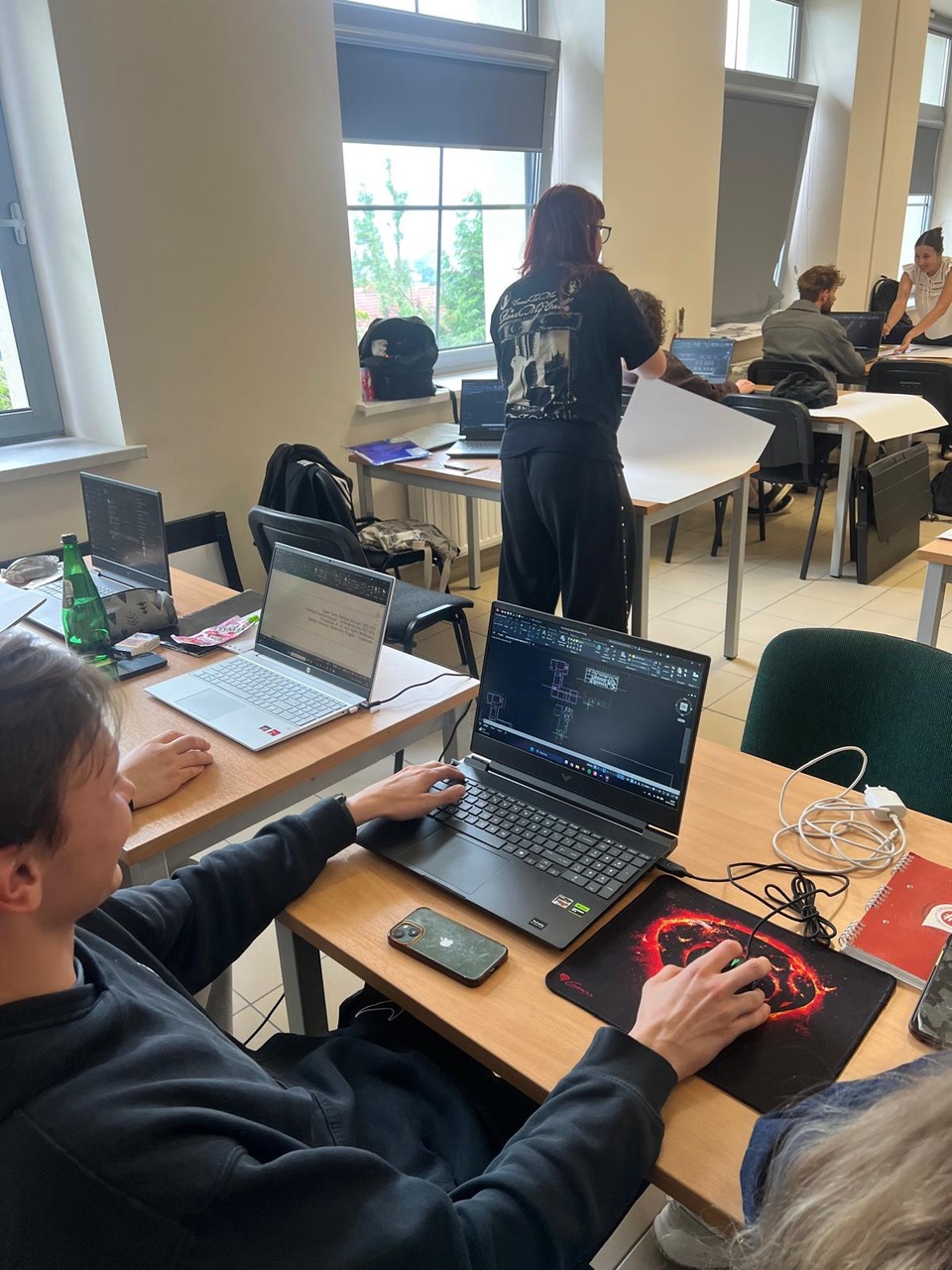 Widok na jasną salę komputerową/klasę, gdzie młody mężczyzna na pierwszym planie operuje myszką, pracując nad rzutami budynku w programie CAD. W tle inne osoby pracują przy swoich stanowiskach.