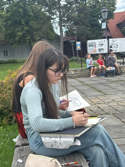 Dziewczyna w okularach i jasnym swetrze skupiona na rysowaniu w szkicowniku. Za nią siedzi kolejna osoba, a w tle widać parkową przestrzeń z ławkami, tablicami informacyjnymi i spacerowiczami.