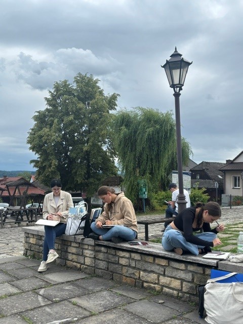 Grupa młodzieży siedząca na kamiennym podwyższeniu pod stylizowaną latarnią, zajęta rysowaniem. W tle rozciąga się widok na drzewa i dachy domów pod pochmurnym niebem.