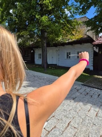 Widok zza ramienia jasnowłosej dziewczyny, która wyciąga rękę przed siebie, trzymając ołówek lub długopis, aby wymierzyć proporcje tradycyjnego, białego drewnianego budynku z ciemnym dachem.