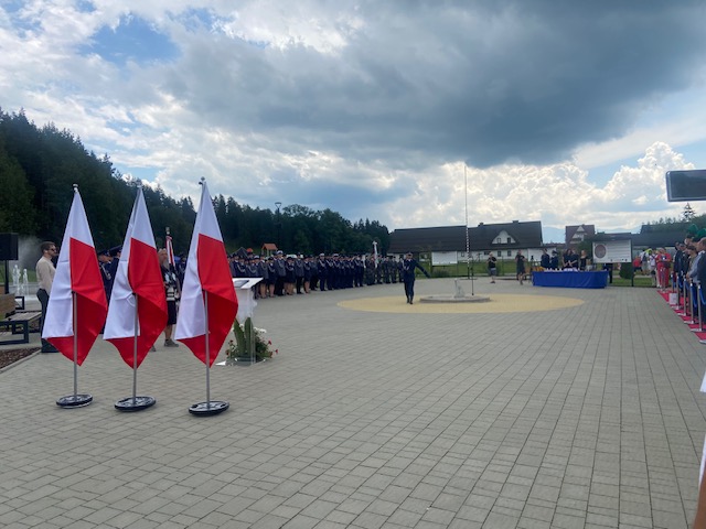 Na pierwszym planie stoją trzy flagi Polski, w tle widać ustawiony w szyku pododdział policjantów oraz spacerującego oficera.