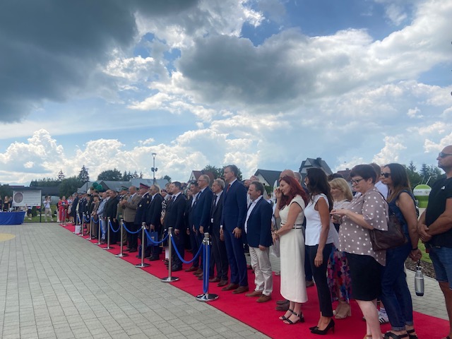 Zgromadzeni goście, urzędnicy i przedstawiciele służb mundurowych stojący wzdłuż czerwonego dywanu, odgrodzeni niebieskim sznurem.