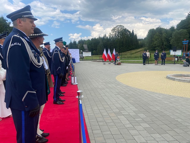 Funkcjonariusze Policji (w tym jeden w tradycyjnym kapeluszu góralskim) oraz inne osoby stojące na czerwonym dywanie podczas trwania uroczystości na placu.