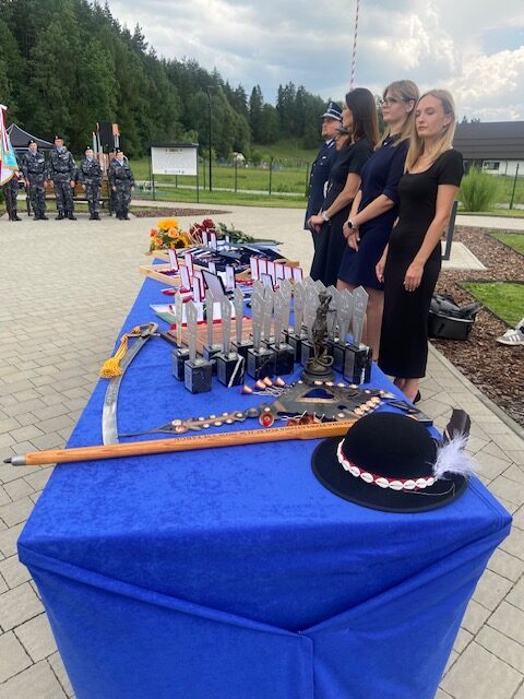 Stół z nagrodami, pucharami, statuetką Temidy, repliką szabli oraz tradycyjnym kapeluszem góralskim. Za stołem stoi kilka osób, w tym kobiety w ciemnych sukienkach oraz policjant.