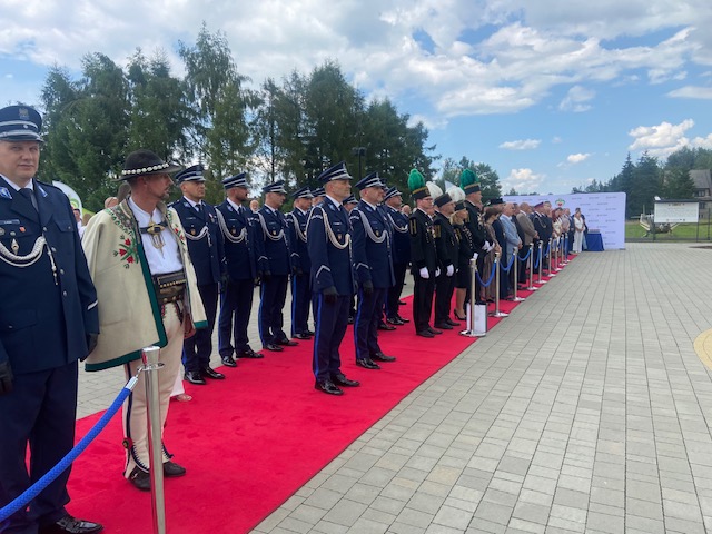 Długi rząd umundurowanych funkcjonariuszy Policji oraz zaproszonych gości stojących na czerwonym dywanie podczas apelu. Jeden z mężczyzn ma na sobie tradycyjny strój podhalański (cuchę).