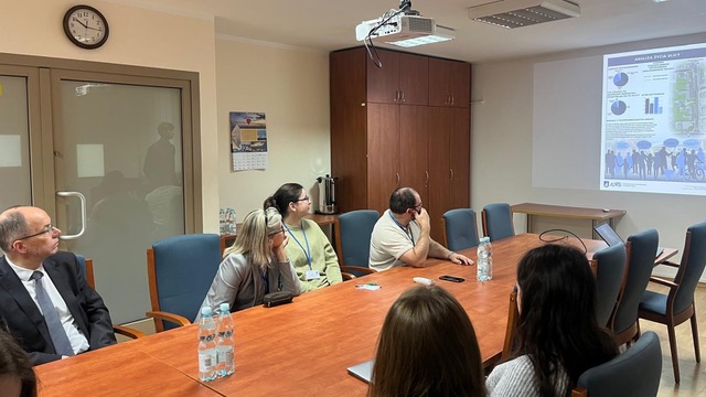 Widok na salę konferencyjną z profilu. Grupa osób siedzi przy dużym, podłużnym stole i ogląda prezentację wyświetlaną na ekranie. Na slajdzie widnieje logo uczelni ANS (Akademia Nauk Stosowanych w Nowym Targu) oraz tytuł "Analiza życia ulicy".