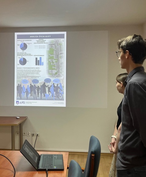 Dwójka młodych ludzi stoi obok ekranu projektora i prezentuje wspomnianą wcześniej "Analizę życia ulicy". Na slajdzie widać wykresy kołowe, słupkowe oraz mapę terenu.