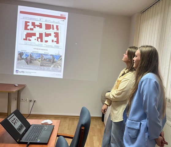 Dwie dziewczyny prezentują kolejny projekt. Na ekranie wyświetlany jest slajd zatytułowany "Otwarcie widokowe", przedstawiający czerwone plany zabudowy oraz zdjęcia perspektywiczne ulic.