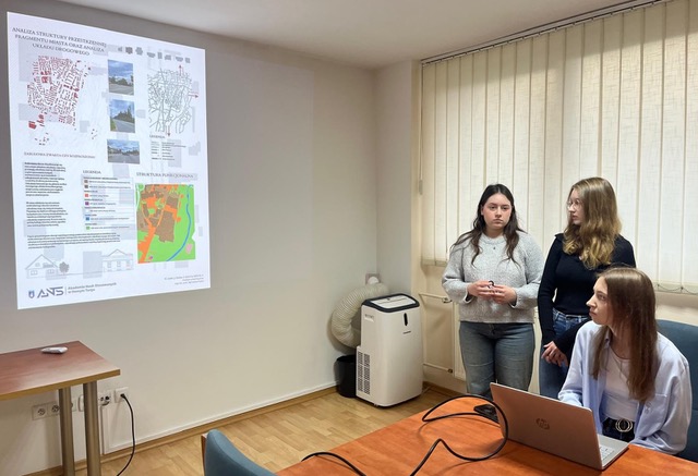 Kolejna grupa trzech dziewcząt (dwie stoją, jedna siedzi przy laptopie) przedstawia projekt pt. "Analiza struktury przestrzennej fragmentu miasta oraz analiza układu drogowego". Na slajdzie widać mapy geodezyjne/urbanistyczne oraz plany funkcjonalne terenu.
