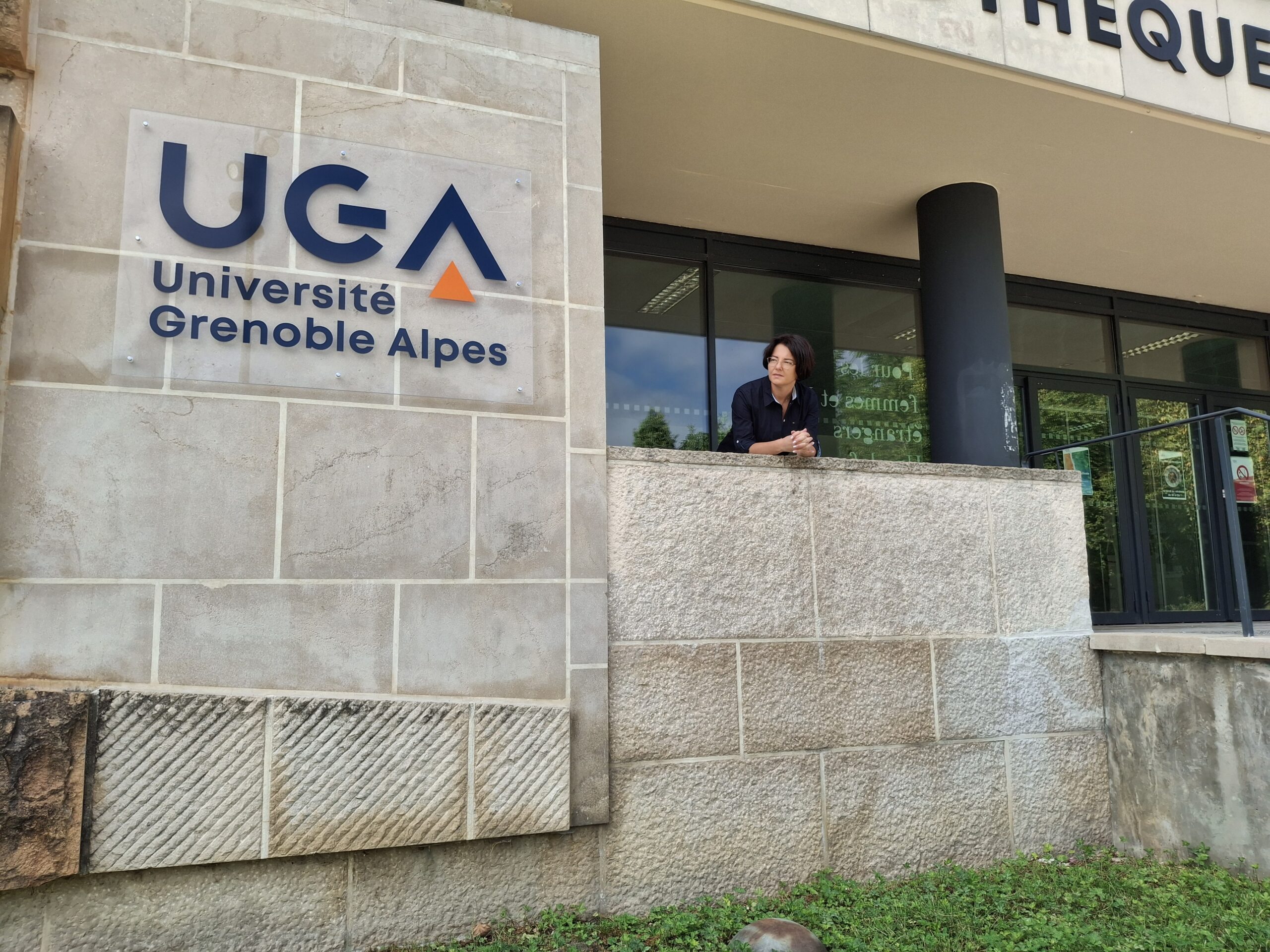 Tablica UGA Université Grenoble Alpes na murze, de Katarzyna Ceklarz wychylająca się z okna obok.