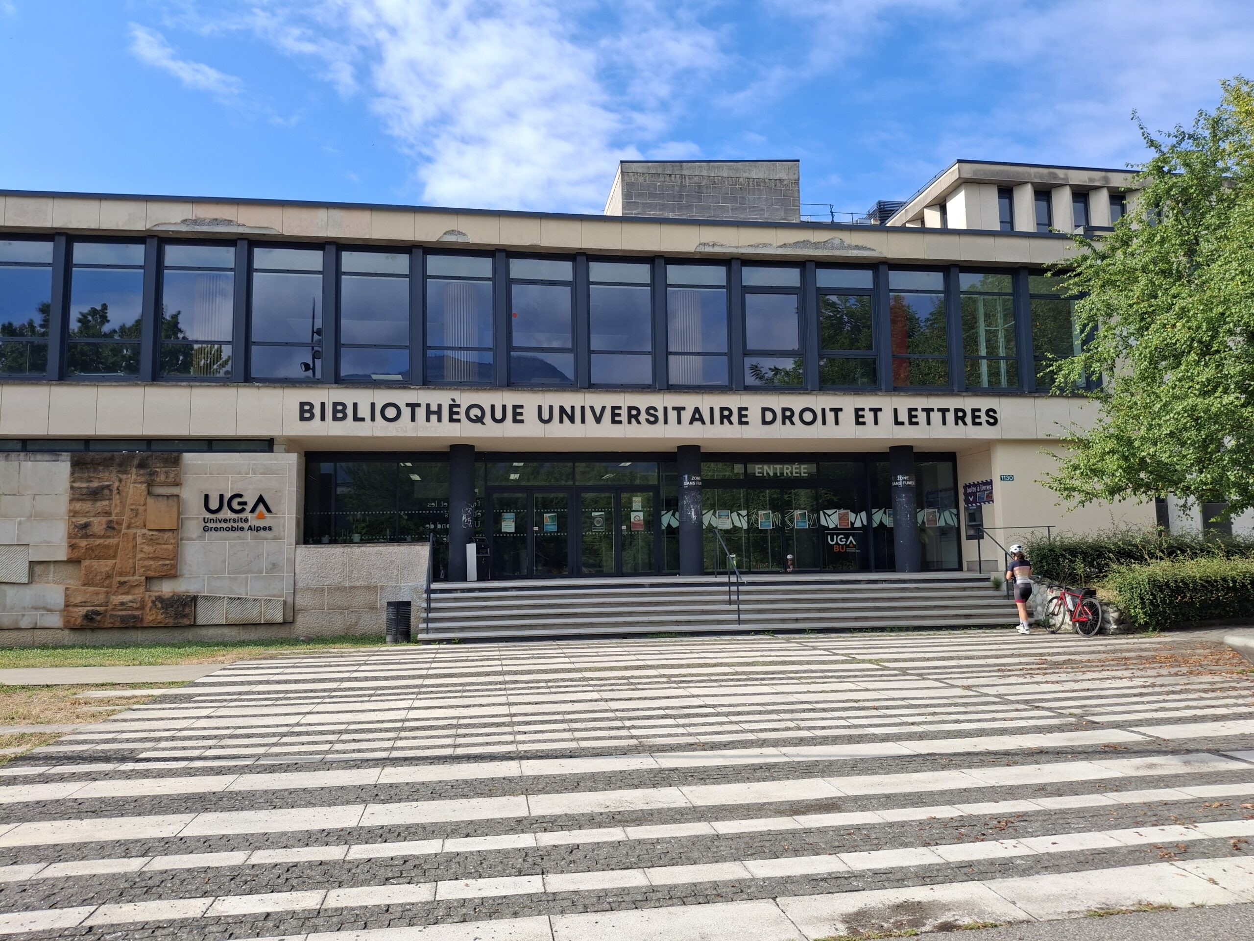 Fasada budynku Bibliothèque Universitaire Droit et Lettres uczelni UGA ze schodami i pasiastym placem z przodu.