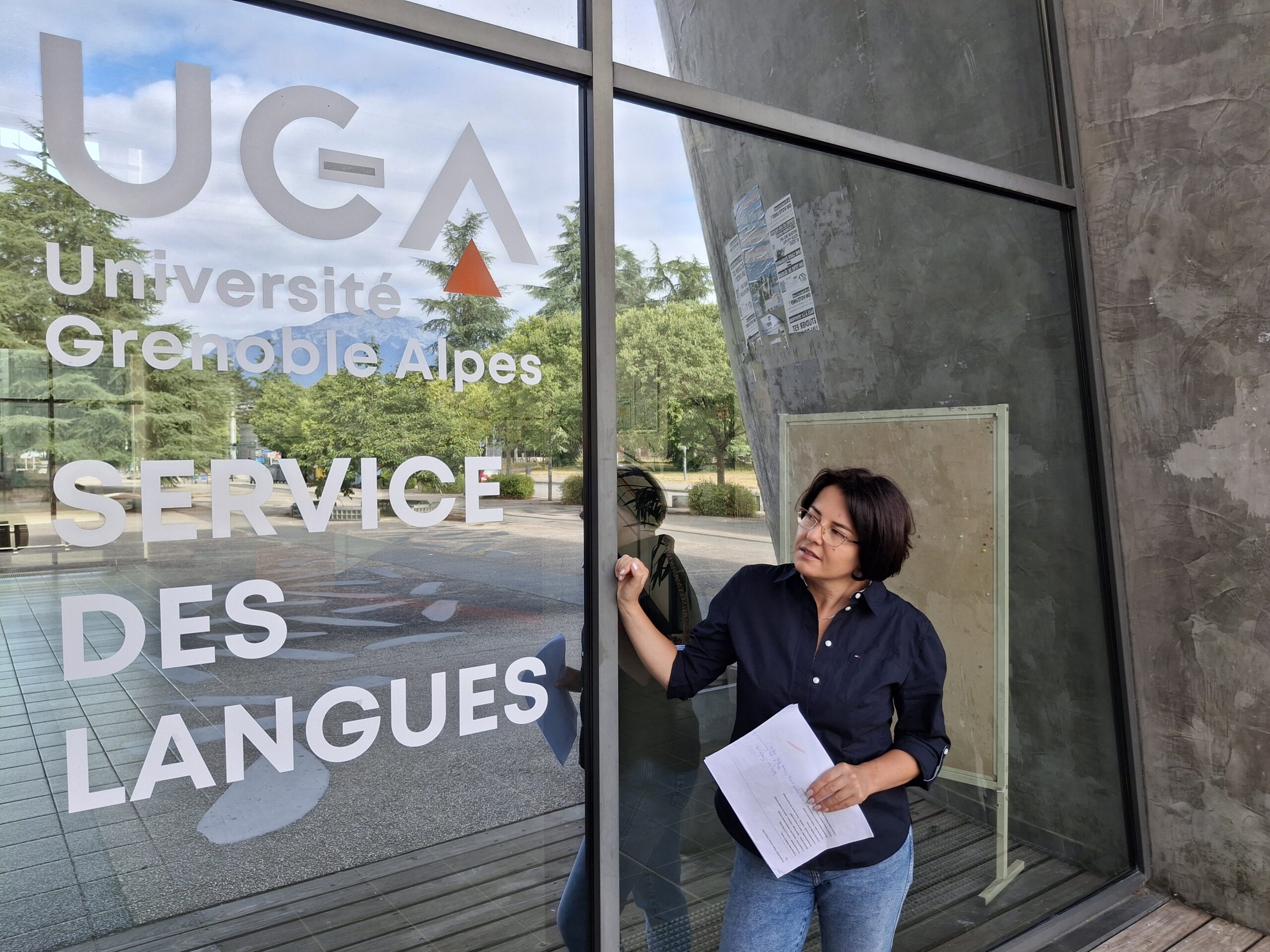 dr Katarzyna Ceklarz z dokumentem w ręku stoi przy przeszklonym wejściu do budynku UGA z napisem Service des Langues.