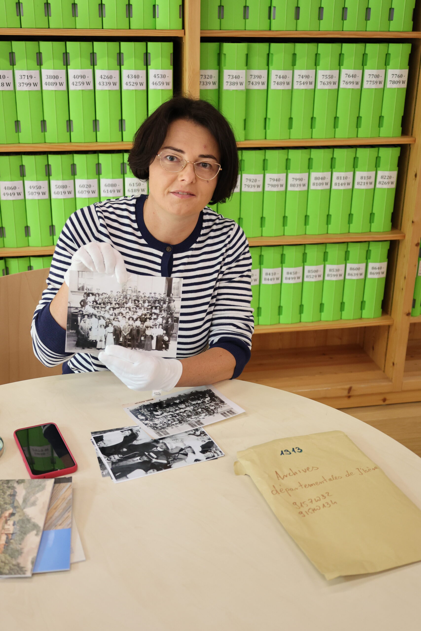 dr Katarzyna Ceklarz w rękawiczkach prezentuje przedwojenną fotografię grupową, obok leży zabytkowa mapa i podpisana koperta, na tle zielonych pudeł archiwalnych.