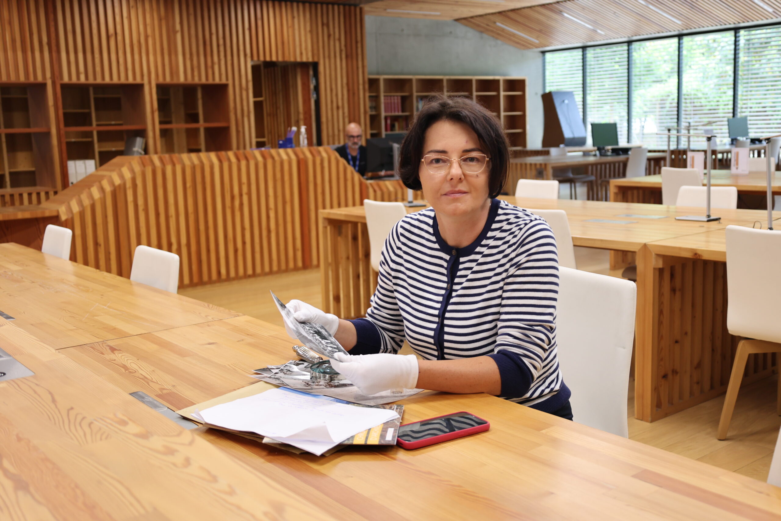 dr Katarzyna Ceklarz w rękawiczkach trzyma starą fotografię przy stole w nowoczesnej czytelni z drewnianymi panelami i biurkami.