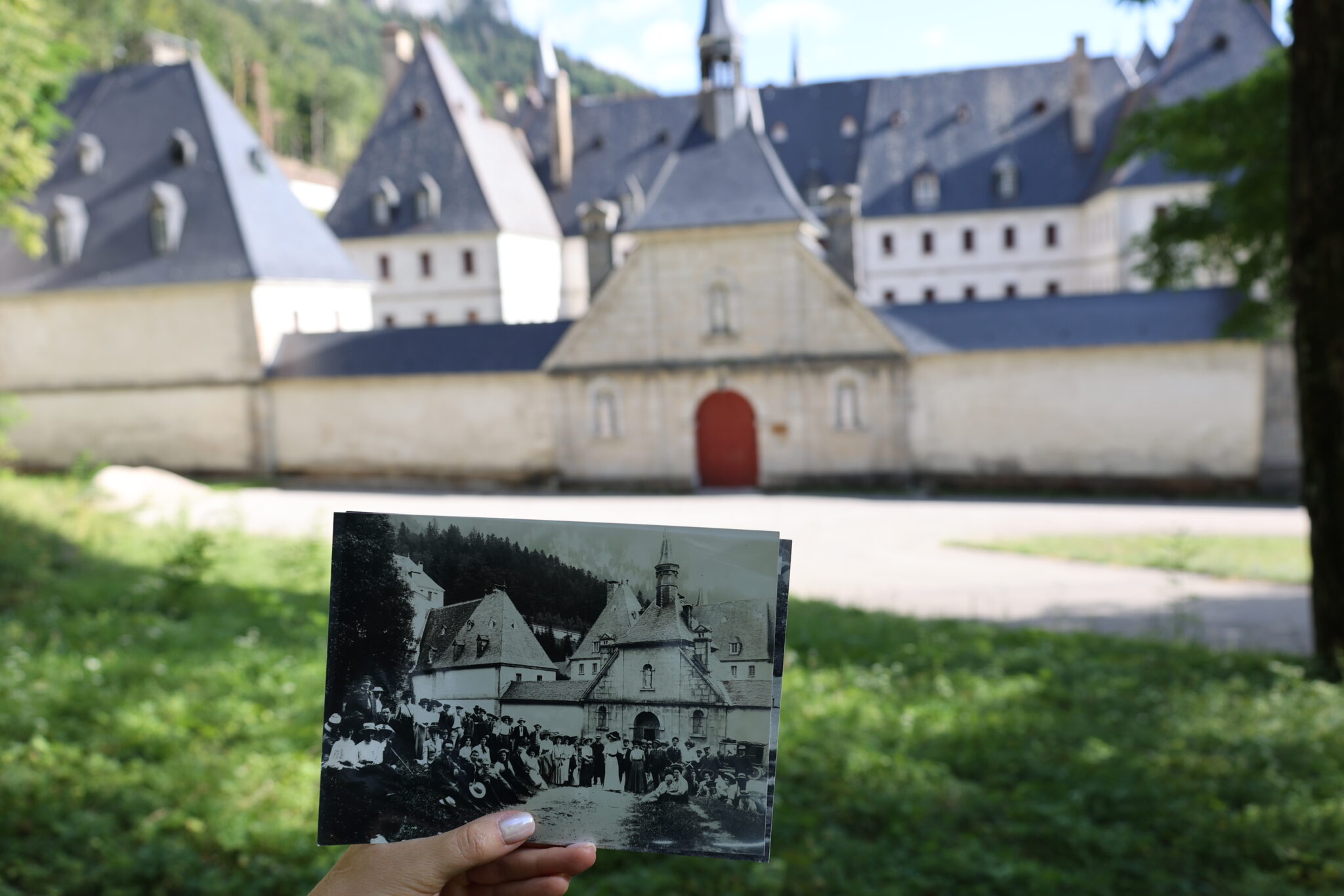 Ręka trzyma starą czarno-białą fotografię na tle współczesnego widoku klasztoru Grande Chartreuse we francuskich Alpach, pokazując przeszłość i teraźniejszość.