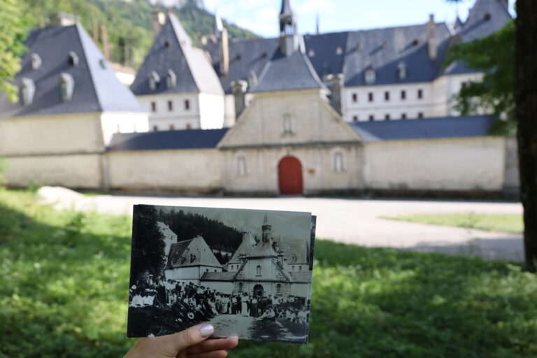 Ręka trzyma starą czarno-białą fotografię na tle współczesnego widoku klasztoru Grande Chartreuse we francuskich Alpach, pokazując przeszłość i teraźniejszość.