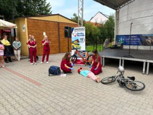 Pokaz pierwszej pomocy przed sceną: studentki medyczne pomagają dziecku leżącemu obok roweru.